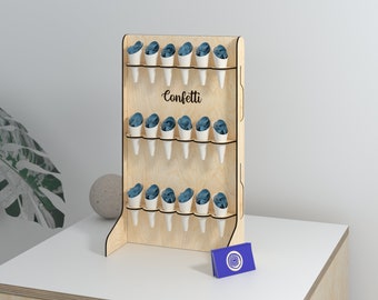 May include: A wooden confetti display stand with multiple rows of white paper cones filled with blue confetti. The word "Confetti" is written in cursive on the stand. A small blue box with a white design sits in front of the stand.