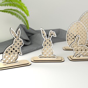May include: Set of five wooden Easter decorations featuring bunnies and eggs. The decorations are made of light brown wood with a white woven pattern. The bunnies are standing on their hind legs with their ears up. The eggs are standing upright.