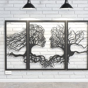 May include: Black metal wall art depicting two faces formed by tree branches, with roots intertwining at the base. The artwork is framed in black and mounted on a white brick wall, with two black pendant lights above.