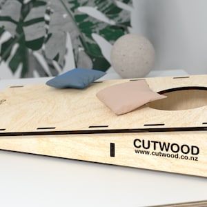 K&ouml;nnte beinhalten: Ein Holz-Beanbag-Wurfspiel mit braunem Sockel und schr&auml;ger Spielfl&auml;che. Das Spiel hat ein kreisrundes Loch in der Mitte der Spielfl&auml;che. Zwei Beanbags sind enthalten, ein blauer und ein beiger.