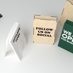 May include: Three wooden A-frame signs with different messages. The first sign is white with black text that reads "Citylake Yoga". The second sign is light brown with black text that reads "Follow us on Social". The third sign is green with white text that reads "We are Open for Business".