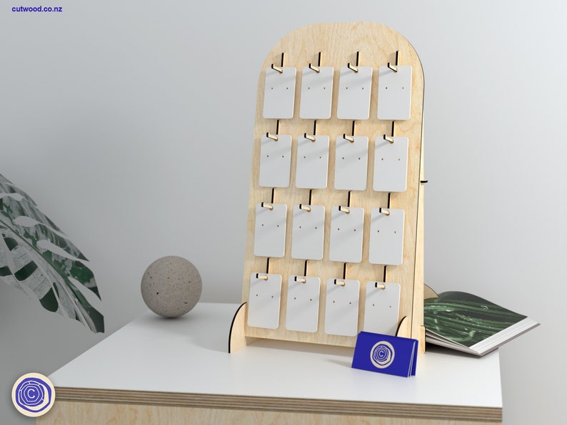 May include: A wooden earring display stand with 20 white card slots for holding earrings. The stand is made of light wood and has a rounded top. The stand is on a light wood table with a green plant and a small grey ball in the background.