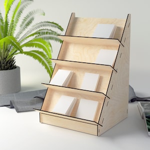 May include: A wooden display stand with four shelves for holding cards or other small items. The stand is made of light wood and has a natural finish. The shelves are angled to create a tiered effect.