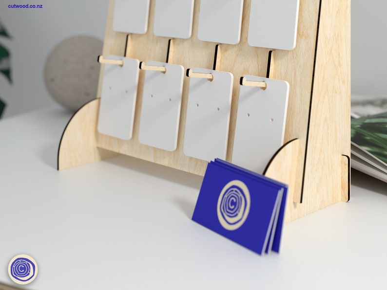 May include: A wooden earring display stand with white card inserts. The stand has a triangular shape and is made of light wood. There are 12 card inserts, each with two holes for displaying earrings. The stand is sitting on a white surface.
