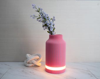 May include: A pink, ribbed vase with a glowing base holds a bouquet of light purple flowers. A white decorative knot sits to the left of the vase. The vase is a modern home decor item.