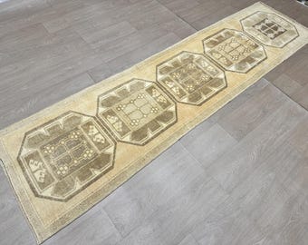 May include: A beige runner rug with a geometric pattern in brown. The rug has four repeating squares with a central design.