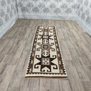 May include: A white and brown patterned runner rug with geometric designs. The rug has a fringe border and is made of a soft, woven material.
