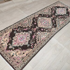 May include: A vintage runner rug with a brown background and a geometric pattern in shades of pink, beige, and blue. The rug has a fringed edge.