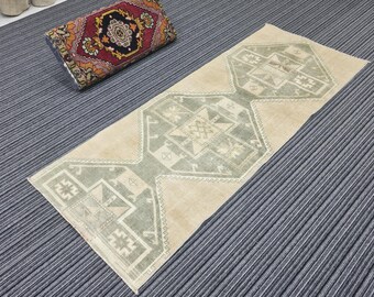 May include: A rectangular beige and green patterned rug with geometric designs. The rug is placed on a gray striped surface. A rolled-up patterned pillow is in the background.