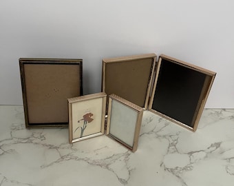May include: A collection of four metallic picture frames with thin, textured borders. One frame is a standalone rectangle, while the other three are connected by hinges, forming a folding display. One small frame contains a print of a red iris flower.
