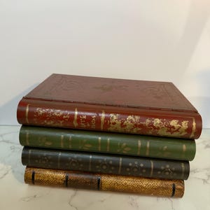 May include: A stack of four decorative books in varying colors. The top book is red with gold accents and a design on the cover. The other books are green, dark brown, and tan, each with gold detailing. The books are stacked on top of each other.