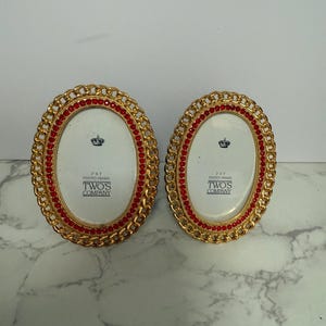 May include: Two oval photo frames with gold-colored frames and red accents. The frames have a chain-like design around the edges and red jewels. The frames have a white interior with the text "TWO'S COMPANY".