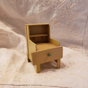 May include: A miniature wooden nightstand painted in a light yellow color. The nightstand has a drawer with a green knob, a shelf, and a small open storage area. The piece is a vintage style, likely for a dollhouse or similar decorative use.