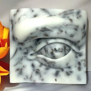 May include: A close-up of a white and gray sculpted eye with a detailed texture. The eye is partially closed, and the eyelid is visible. The sculpture is made of a smooth, white material with a gray marbling effect.