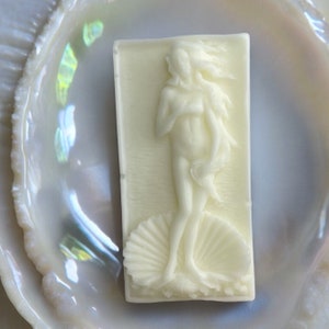 Puede incluir: Pastilla de jabón blanca con una imagen en relieve de una mujer de pie sobre una concha. La mujer está desnuda y tiene el pelo largo y suelto.
