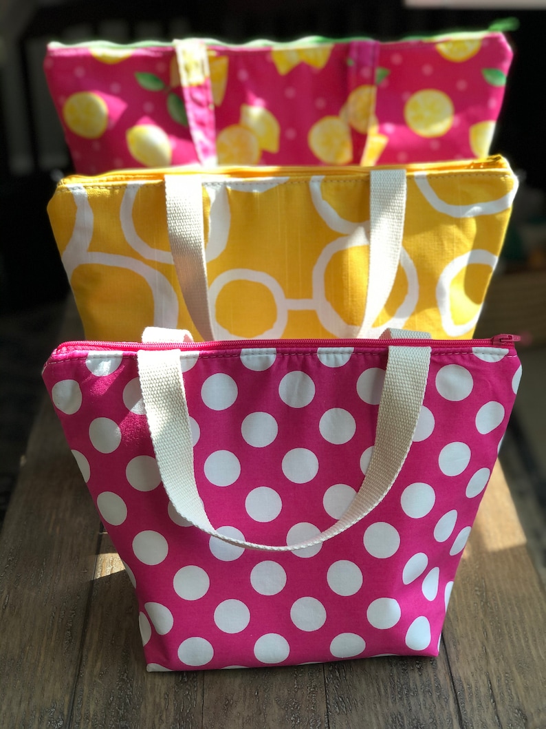 Peut inclure: Trois sacs isothermes r&eacute;utilisables avec fermeture &eacute;clair et poign&eacute;es blanches. Le sac du haut a un fond rose avec des citrons et des feuilles vertes. Le sac du milieu a un fond jaune avec des cercles blancs. Le sac du bas a un fond rose avec des pois blancs.