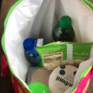Peut inclure: Un sac &agrave; d&eacute;jeuner isotherme rose et jaune avec une fermeture &eacute;clair verte. Le sac est ouvert et contient un r&eacute;cipient en plastique transparent avec un couvercle vert, un r&eacute;cipient en plastique transparent avec une substance brune, une brique de jus bleue et blanche, un r&eacute;cipient en plastique vert avec un couvercle bleu, une bouteille en verre transparente avec un couvercle vert et un sac de chips avec une &eacute;tiquette de valeur nutritionnelle. Le sac de chips porte le texte "Love our chips? Give us a call 1-877-VICKI-CALL" et "Miss Vicki" imprim&eacute; dessus.