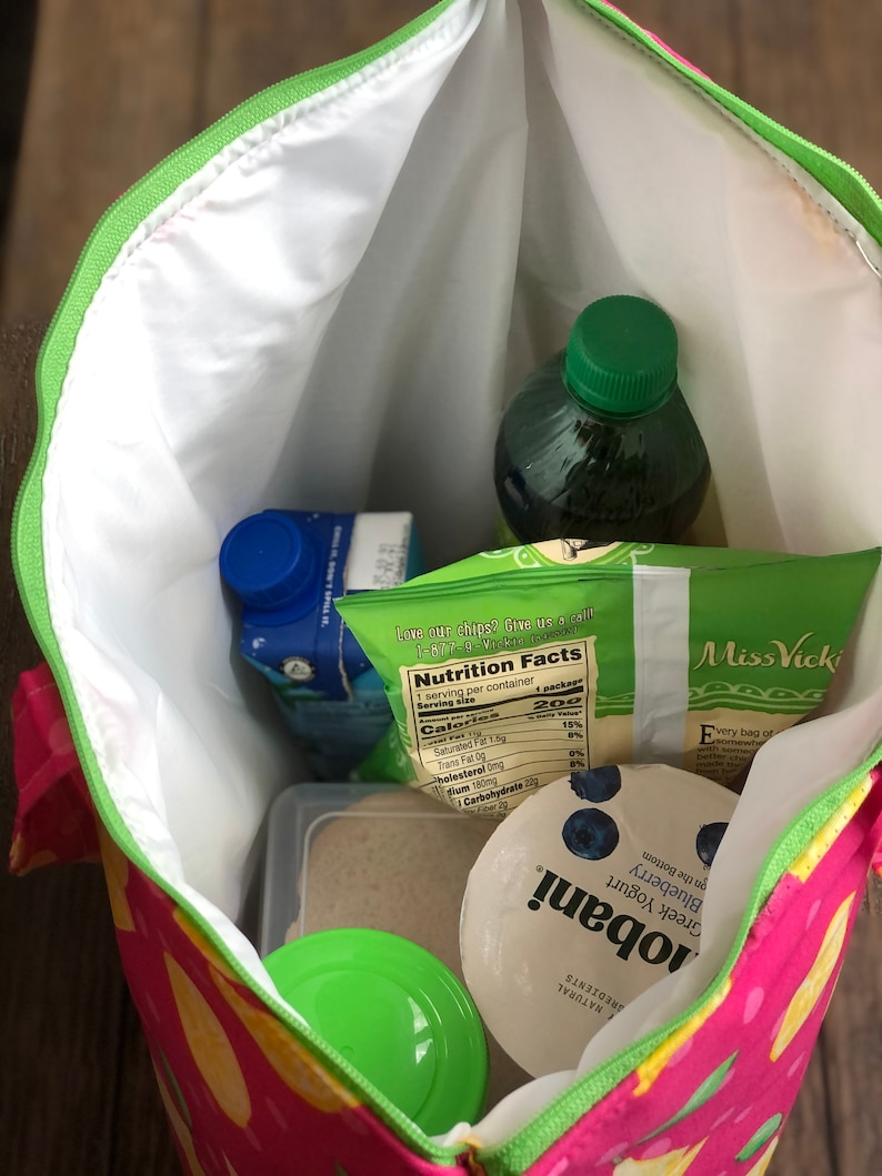 Peut inclure: Un sac &agrave; d&eacute;jeuner isotherme rose et jaune avec une fermeture &eacute;clair verte. Le sac est ouvert et contient un r&eacute;cipient en plastique transparent avec un couvercle vert, un r&eacute;cipient en plastique transparent avec une substance brune, une brique de jus bleue et blanche, un r&eacute;cipient en plastique vert avec un couvercle bleu, une bouteille en verre transparente avec un couvercle vert et un sac de chips avec une &eacute;tiquette de valeur nutritionnelle. Le sac de chips porte le texte "Love our chips? Give us a call 1-877-VICKI-CALL" et "Miss Vicki" imprim&eacute; dessus.