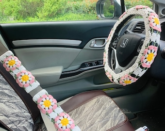 May include: A crocheted steering wheel cover and seatbelt cover with pink and white daisy designs. The covers are made of a soft, textured yarn.
