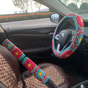 Peut inclure: L'intérieur d'une voiture avec un couvre-volant en crochet et un couvre-ceinture de sécurité en crochet. Le couvre-volant est bleu, rose et blanc avec un motif floral. Le couvre-ceinture de sécurité est rose, bleu et blanc avec un motif floral.