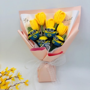 Puede incluir: Un ramo de tulipanes amarillos de crochet con personajes de minions de crochet unidos a los tallos. El ramo está envuelto en papel de seda rosa y atado con una cinta blanca.