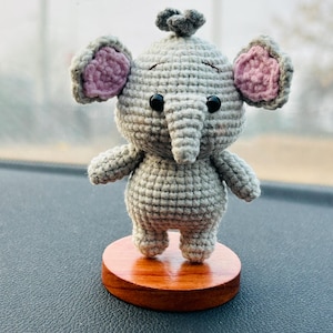 May include: A handmade crochet elephant figurine in gray with pink inner ears and black eyes. The elephant is standing on a small, round, wooden base. The trunk is pointing downwards.