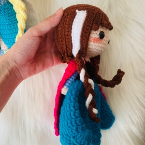Puede incluir: Una mu&ntilde;eca de crochet con cabello casta&ntilde;o, una trenza blanca y un vestido azul y rosa. La mu&ntilde;eca sostiene una trenza blanca en su mano.