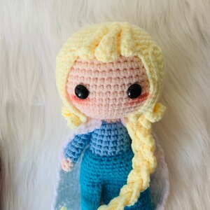 Puede incluir: Una mu&ntilde;eca de crochet con el pelo largo rubio, ropa azul y azul claro y ojos negros.