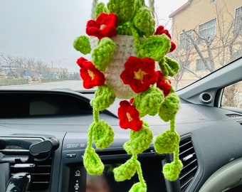 Op de afbeelding: Een gehaakte hangende plantenpot met rode bloemen en groene bladeren. De pot hangt aan een achteruitkijkspiegel in een auto.