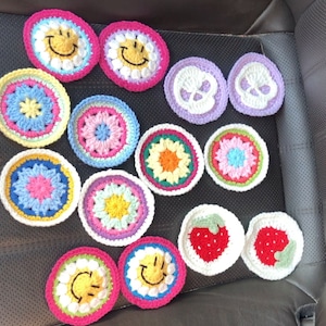 May include: A collection of colorful, handmade crochet coasters. Designs include smiley faces, floral patterns, skulls, and strawberries. The coasters are made with various colors of yarn, including pink, yellow, green, blue, and white.