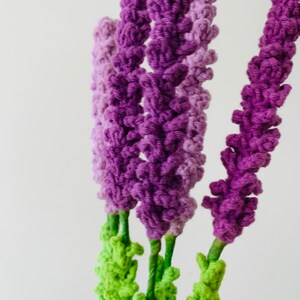 May include: A bunch of purple and green crocheted flowers. The flowers are made of yarn and have a textured, fluffy appearance.
