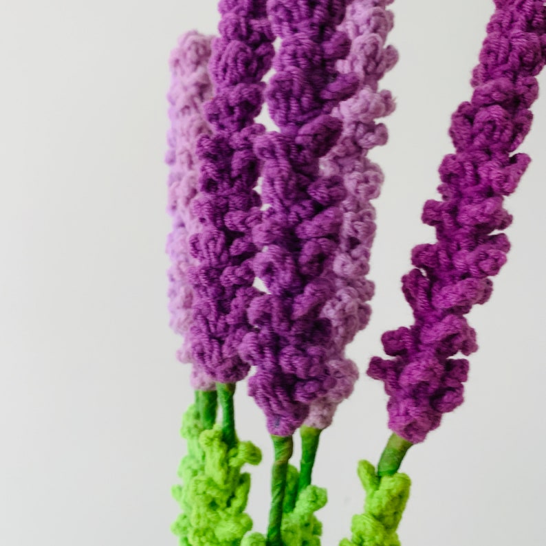 May include: A bunch of purple and green crocheted flowers. The flowers are made of yarn and have a textured, fluffy appearance.