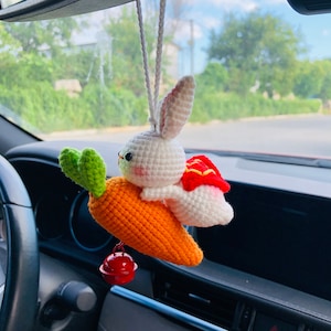 May include: A white crocheted bunny rabbit wearing a red crocheted cape sits on a crocheted orange carrot. The carrot has green leaves and a small red bell hanging from the bottom. The bunny and carrot are hanging from a rearview mirror in a car.