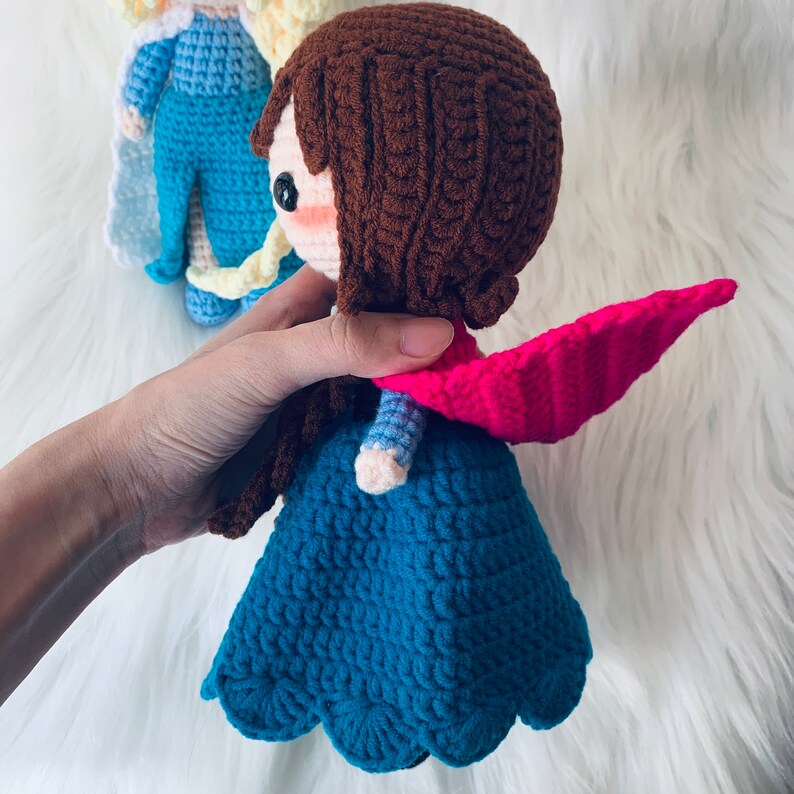 Puede incluir: Una mu&ntilde;eca de crochet con cabello casta&ntilde;o, un vestido azul y una capa rosa. La mu&ntilde;eca sostiene una mu&ntilde;eca de crochet azul y blanca en su mano.