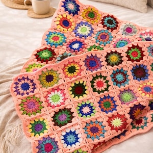 May include: A colorful, handmade crochet blanket with a floral pattern. The blanket features a peach-colored base with vibrant, square floral motifs in various colors, including blue, green, yellow, and pink. The blanket is folded on a bed.