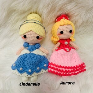 Peut inclure: Deux poup&eacute;es princesses en crochet, l'une en robe bleue avec une couronne blanche et l'autre en robe rose avec une couronne rouge. Les poup&eacute;es sont sur une surface blanche et moelleuse. Le texte "Cendrillon" est sous la poup&eacute;e bleue et "Aurore" est sous la poup&eacute;e rose.