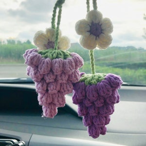 Op de afbeelding: Twee gebreide druiventrossen met groene stelen en witte bloemen die aan een achteruitkijkspiegel hangen. De druiven zijn lichtpaars.