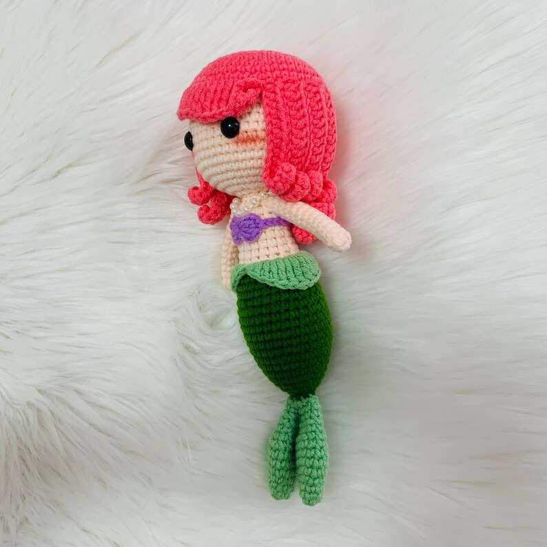 Puede incluir: Una mu&ntilde;eca de sirena de crochet con pelo rosa, un top morado y blanco y una cola verde.