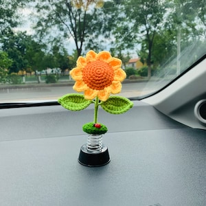 May include: A crocheted sunflower with green leaves and a red ladybug on a spring-loaded base. The sunflower is yellow and orange and is sitting on a black base.