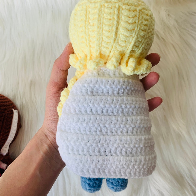 Puede incluir: Una mu&ntilde;eca de crochet con pelo amarillo, un vestido blanco y pantalones azules. La mu&ntilde;eca est&aacute; hecha a mano y tiene un aspecto dulce y caprichoso.