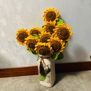 May include: A bouquet of crocheted sunflowers in a white vase with a peacock design. The sunflowers are yellow and green and the vase has a floral pattern.
