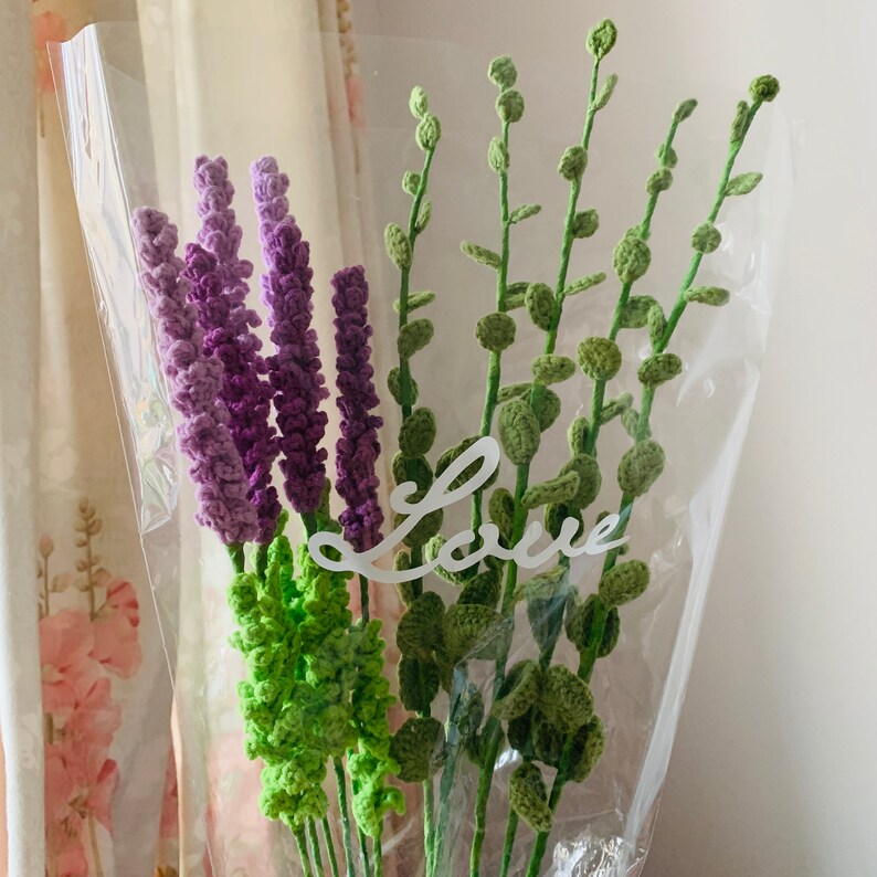 May include: A bouquet of crocheted lavender and green foliage flowers in a clear plastic wrap with the word "Love" written on it.