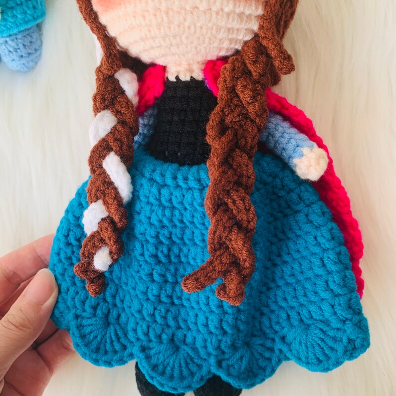 Puede incluir: Mu&ntilde;eca de crochet con cabello casta&ntilde;o, un vestido azul y una capa rosa. La mu&ntilde;eca tiene un peinado trenzado blanco y marr&oacute;n.