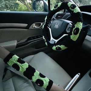 May include: A black and green crocheted steering wheel cover with a pattern of cartoon eyes. A matching black and green crocheted seatbelt cover is also visible.