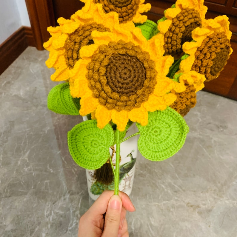 May include: A bouquet of crocheted sunflowers in yellow and brown with green leaves. The sunflowers are arranged in a vase with a peacock design.