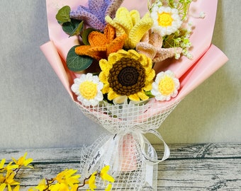 Puede incluir: Un ramo de flores de ganchillo en un envoltorio de papel rosa y blanco. El ramo incluye un girasol, una flor morada, una flor amarilla y una flor rosa. El ramo está atado con una cinta blanca.