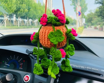 Op de afbeelding: Een gehaakte hangplant met groene bladeren en roze bloemen in een bruine pot. De plant hangt aan een achteruitkijkspiegel in een auto.