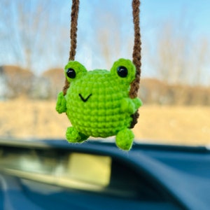 Op de afbeelding: Een groene gehaakte kikker die aan een bruin touwtje hangt. De kikker heeft zwarte ogen en een kleine glimlach.