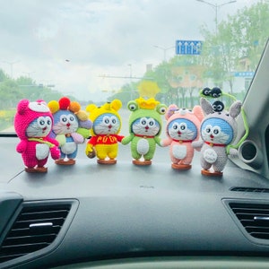 May include: Seven crocheted plush toys in various colors and designs, including pink, blue, green, and yellow, are displayed on a car dashboard. The toys are all wearing different outfits and have different facial expressions.