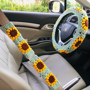 以下が含まれることがあります： 車の中の、かぎ針編みで作ったひまわり柄のハンドルカバーとシートベルトカバー。ハンドルカバーは緑色で、中心が茶色、花びらが黄色。シートベルトカバーは緑色で、中心が茶色、花びらが黄色です。